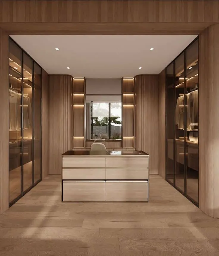 A dressing room lined in pale oak, with glass-fronted wardrobes down both walls and a drawer island under the window