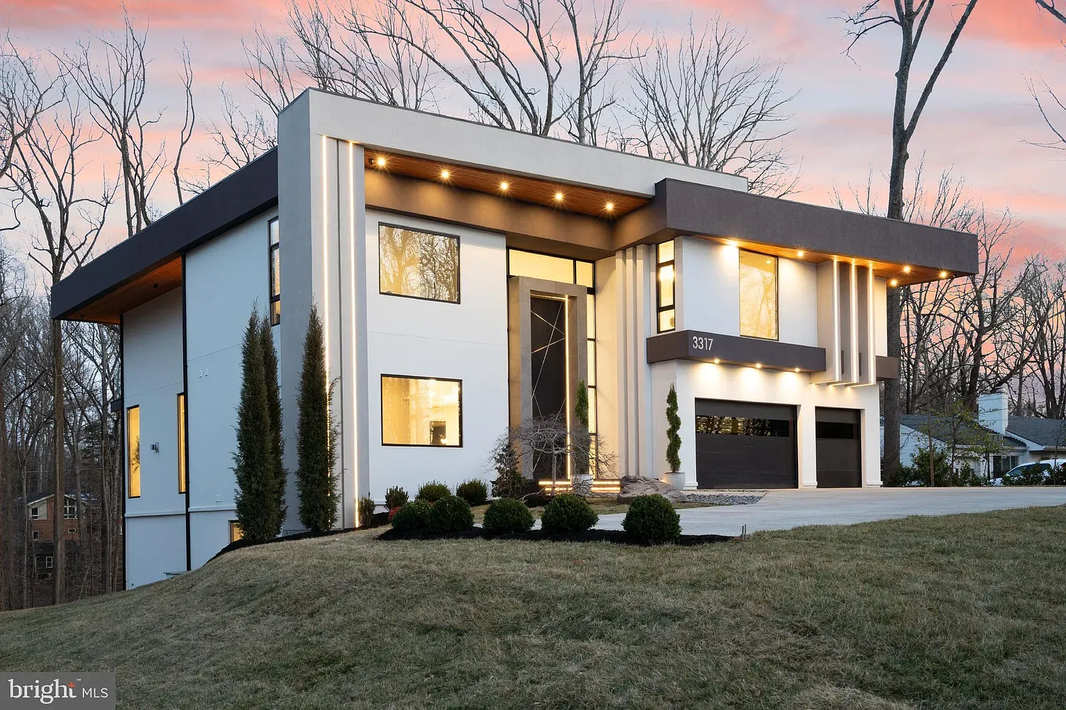 The house at dusk in winter — a flat-roofed modern facade in white stucco, vertical light strips down its corners and a lit wood soffit over the entrance
